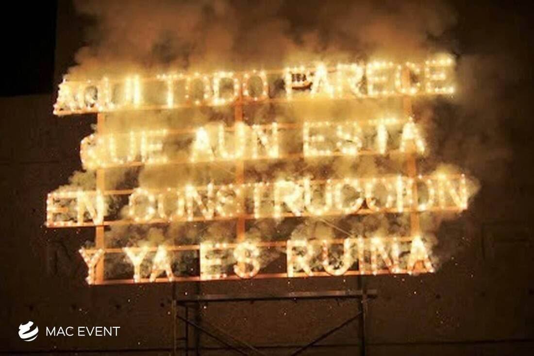 A large firework sign with Spanish text "Aquí todo parece que aún está en construcción y ya es ruina" ("Here everything still seems under construction, and it’s already a ruin"), glowing with golden light.